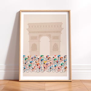This photo of a contemporary cycling art print in ecru, features a colourful peloton riding past the Arc de Triomphe in Paris. The print is framed in a simple oak frame and is sat on a wooden herringbone style floor and leaning against a white wall.
