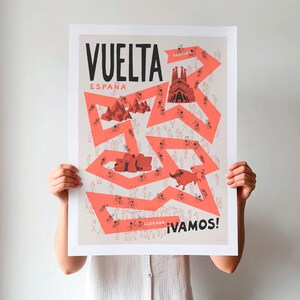Person holding an unframed Vuelta Grand Tour cycling print with a red illustrated cycling route around Spain, including playful illustrations of cyclists, fans and famous Spanish landmarks against a neutral background.