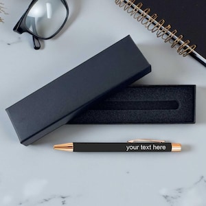 May include: A black pen with gold detailing and the text "your text here" is next to an open black pen box. A black notebook, glasses, and decorative flowers are also in the image.