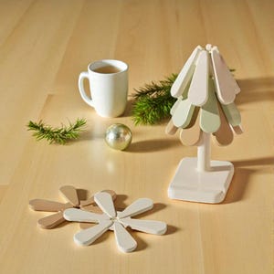 Puede incluir: Un árbol de Navidad decorativo con base blanca y elementos en forma de pétalo en capas en tonos de blanco, verde y marrón. También hay dos posavasos a juego y una taza blanca, junto con un adorno plateado y ramitas de vegetación.