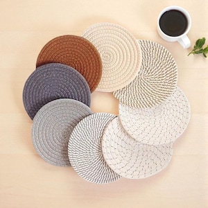 May include: A collection of round coasters in various neutral colors, including brown, gray, and cream. The coasters are made of woven material and arranged in a circular pattern. A white mug filled with coffee sits nearby.