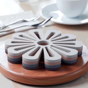 Puede incluir: Un salvamanteles con forma de flor, con un diseño en capas en tonos grises, rosas y marrones. El salvamanteles está sobre un posavasos redondo de madera. Protege las superficies de ollas y sartenes calientes, con un diámetro de unos 18 cm.