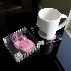 May include: A white ceramic mug with a face design sits on a clear square coaster. Stacked clear coasters with a pink and blue heart diagram are in the foreground. The coasters have the text "CARDIOLOGY DEPT. NO. 1".