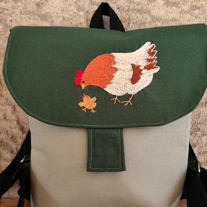 Pode incluir: Mochila verde e bege com um desenho bordado de galinha e pintinho. A aba é verde escuro e o corpo principal é bege. A galinha é laranja, branca e castanha, com uma crista vermelha. O pintinho é amarelo.