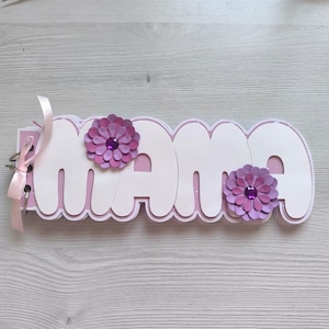 May include: A handmade scrapbook with the word "Mama" in white, layered letters. Two purple paper flowers with jewel centers adorn the letters. A pink ribbon is tied to the binding. The background is a light wood grain.