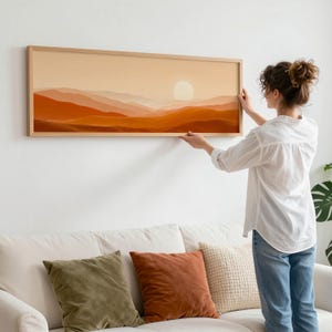 May include: A rectangular framed artwork depicting a desert landscape with a setting sun. The artwork features shades of orange and brown, with a light-colored sun in the center. The frame is made of light-colored wood.