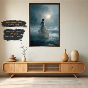 Op de afbeelding: Een ingelijste canvas print met een vrouw in een witte jurk in de oceaan onder een volle maan. Het kunstwerk is in een donkere, sfeervolle stijl. De tekst op het canvas luidt "Brown Framed Canvas Ready to Hang" en "100% Cotton Canvas (300 g/m&sup2;) Premium Print Quality."