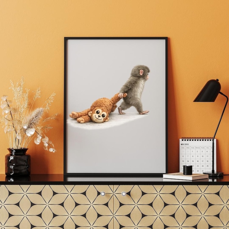 May include: A framed art print showing a small, grey monkey pulling a brown stuffed monkey. The image is set against a light grey background. The print is in a black frame, creating a playful and whimsical scene.