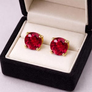 Könnte beinhalten: Ein Paar Ohrstecker mit großen, runden, facettierten roten Edelsteinen, die in goldfarbenem Metall gefasst sind. Die Ohrringe werden in einer offenen schwarzen Schmuckschatulle mit weißem Innenraum präsentiert.