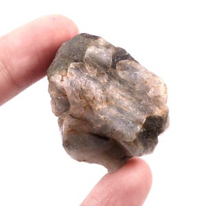 May include: A rough, uncut gemstone held between two fingers. The stone has a muted gray-green color with darker patches and a textured surface. The overall shape is irregular, with visible ridges and variations in tone, suggesting a natural origin.