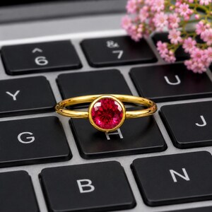 May include: A gold ring featuring a round, faceted, vibrant pinkish-red gemstone set in a bezel setting. The ring is positioned on a black computer keyboard with white lettering, with small pink flowers visible in the upper right corner.