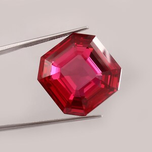 Könnte beinhalten: Ein leuchtend tiefroter Edelstein mit facettiertem Achteckschliff. Der Stein weist eine hohe Klarheit und Lichtreflexion auf seinen polierten Oberflächen auf und wird von einer silberfarbenen Metallpinzette vor neutralem Hintergrund gehalten.