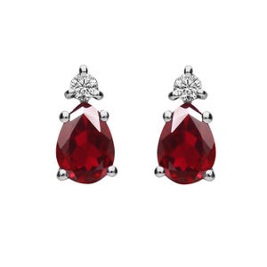 May include: A pair of stud earrings featuring pear-shaped, deep red gemstones. Each earring has a small, round, sparkling white stone at the top and silver-colored metal accents. The earrings are set against a white background.