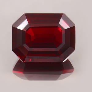 May include: A rectangular-cut gemstone with a rich, deep red colour. The facets of the gem catch the light, producing a sparkling effect. The gem is displayed against a light background, with its reflection visible.