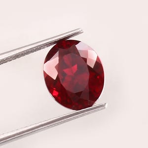 May include: A deep red, oval-cut gemstone held by silver-toned metal tweezers. The stone features a faceted surface that reflects light, showcasing a rich, saturated crimson color throughout its transparent, polished structure.