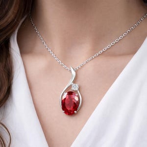 Puede incluir: Un collar de tono plateado con un colgante que presenta una gran piedra preciosa de color rojo intenso con corte ovalado y una pequeña piedra clara de corte redondo encima. El colgante cuelga de una fina cadena plateada sobre tela blanca.