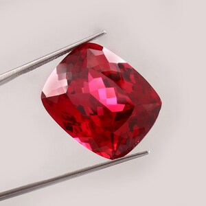 Puede incluir: Una gema roja de talla cojín sostenida por unas pinzas de metal plateado. La piedra presenta una superficie facetada que refleja la luz, creando reflejos de color carmesí intenso y escarlata brillante en su estructura cristalina.