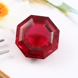 Puede incluir: Una gema de rubí facetada, octogonal, de color rojo intenso. La gema se muestra sobre una superficie blanca, con unas pinzas plateadas en el fondo. Las facetas de la gema reflejan la luz, mostrando su claridad.