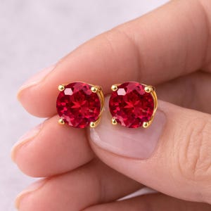 May include: A pair of round-cut, vibrant red gemstone stud earrings set in polished yellow gold. Each earring features a faceted stone secured by four small gold prongs, creating a classic circular design that reflects light across the surface.
