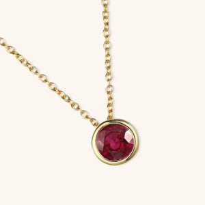 Peut inclure: Un collier à chaîne en or avec une pierre précieuse rubis rouge foncé, ronde et facettée, sertie dans une monture en or lisse et polie. Le pendentif est suspendu à une fine chaîne en or à maillons circulaires uniformes.