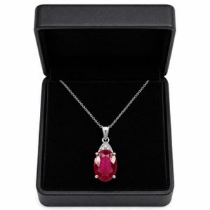 May include: A silver necklace featuring an oval-cut deep red ruby pendant suspended from a delicate chain. The pendant is accented with small clear stones at the top and is presented inside a black velvet-lined jewelry box with a hinged lid.