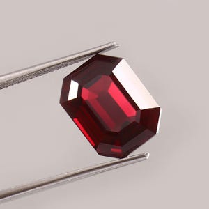 Op de afbeelding: Een dieprode edelsteen in smaragdgeslepen vorm, vastgehouden door een zilverkleurige metalen pincet. De steen heeft een rechthoekige vorm met gefacetteerde randen die het licht reflecteren en een rijke, verzadigde karmozijnrode kleur tonen.