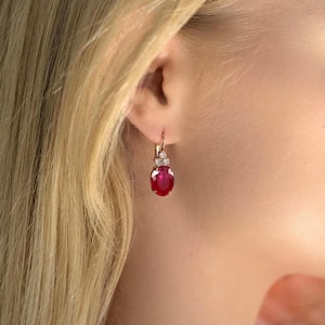 Puede incluir: Un pendiente colgante de oro con una vibrante piedra preciosa roja de forma ovalada suspendida de un pequeño elemento decorativo adornado con tres diminutas piedras claras. El pendiente se asegura con un cierre de palanca de metal pulido.