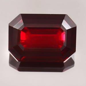 May include: A faceted, rectangular-cut, deep red gemstone. The gem has a reflective surface and is set against a light background. The cut of the gem is an emerald cut, with a series of facets that create a brilliant display of light.