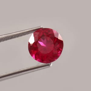 Op de afbeelding: Een ronde, gefacetteerde robijn edelsteen, vastgehouden door een zilveren pincet. De edelsteen heeft een diepe, levendige rode kleur, met meerdere facetten die het licht vangen. De achtergrond is effen lichtgrijs.