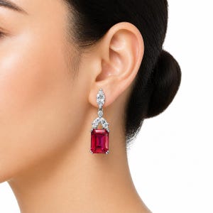 May include: A drop earring featuring a large, rectangular, deep red gemstone suspended from a silver-toned setting. The setting includes a marquise-cut clear stone at the top and two smaller marquise-cut clear stones above the red gemstone.