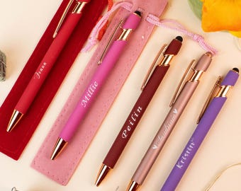 May include: A collection of personalized pens in various colors, including red, pink, and purple, each with a gold-colored tip and clip. The pens are displayed with matching velvet pouches, and each pen is engraved with a name. The pens are a gift item.