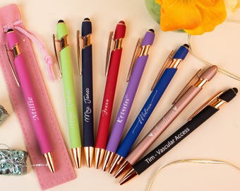 May include: A collection of personalized pens in various colors, including pink, green, navy, red, purple, blue, and rose gold. Each pen has a gold-colored clip and tip, with names or phrases engraved on them. A pink pen case is also present.