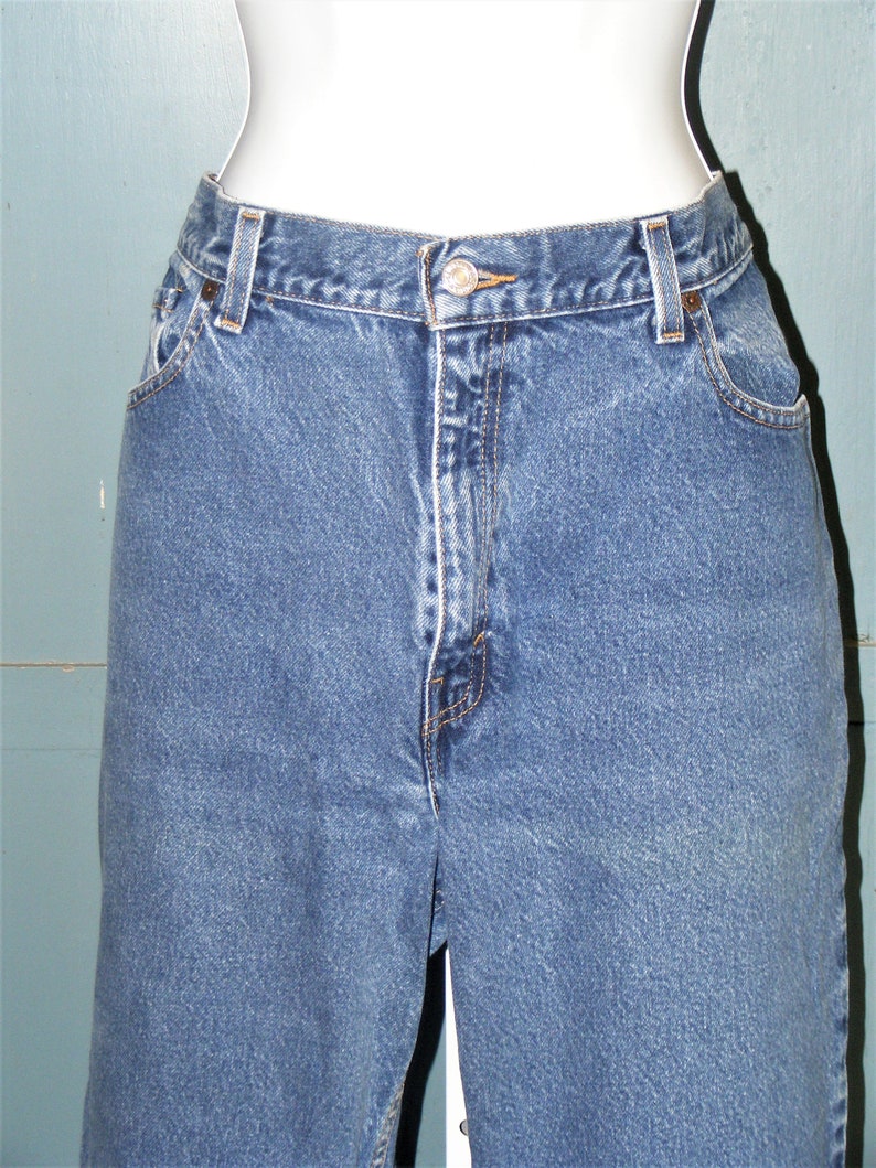 May include: Close-up of blue denim jeans. The jeans have a button closure, belt loops, and pockets. The denim is a medium wash with visible stitching details. The jeans are displayed on a mannequin.