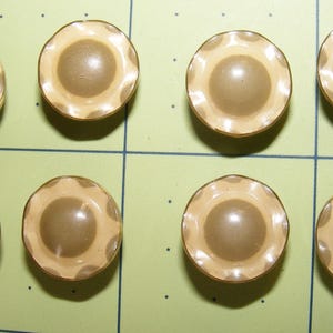 Könnte beinhalten: Acht Vintage-Knöpfe mit einem wellenförmigen Randdesign. Die Knöpfe haben einen hellbraunen Außenring und eine dunklere braune Mitte. Sie sind auf einer hellgrünen Oberfläche mit Rasterlinien angeordnet.
