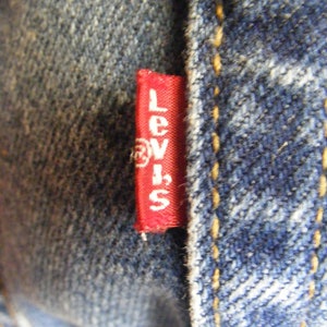 May include: Close-up of denim fabric with a red tag featuring the brand name "Levi's" in white lettering. The tag is sewn into the blue denim, with visible yellow stitching.