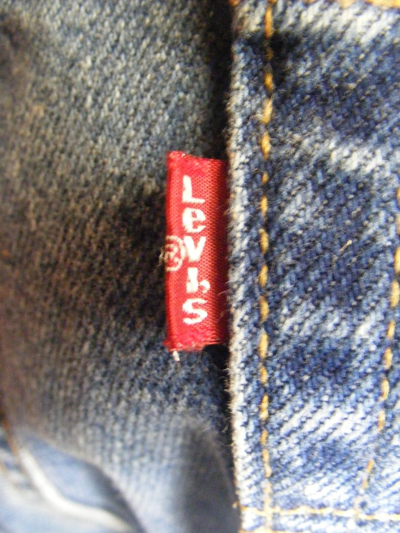May include: Close-up of denim fabric with a red tag featuring the brand name "Levi's" in white lettering. The tag is sewn into the blue denim, with visible yellow stitching.
