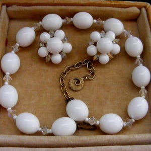 May include: A vintage white beaded necklace and matching earrings set. The necklace features large, oval beads interspersed with smaller, clear crystal beads. The earrings are clusters of small, round white beads. The set is displayed in a tan jewelry box.