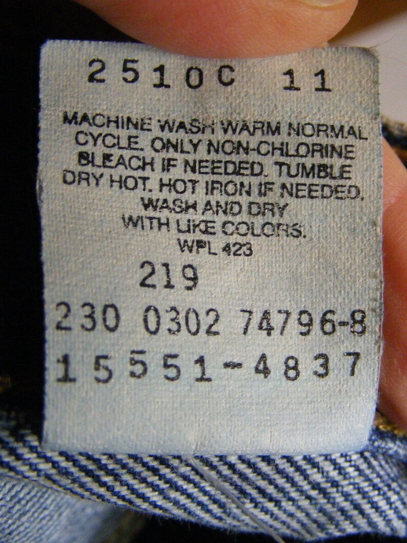 May include: A close-up of a white clothing tag with black text. The tag includes care instructions: machine wash warm, non-chlorine bleach if needed, tumble dry hot, and hot iron if needed. The tag also has several numerical codes.