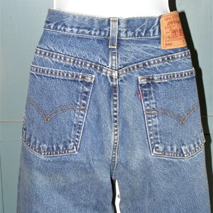 May include: Close-up of the back of blue denim jeans. The jeans have two back pockets with a classic curved design, a small red tag, and a brown leather Levi Strauss label. The jeans are displayed on a mannequin.
