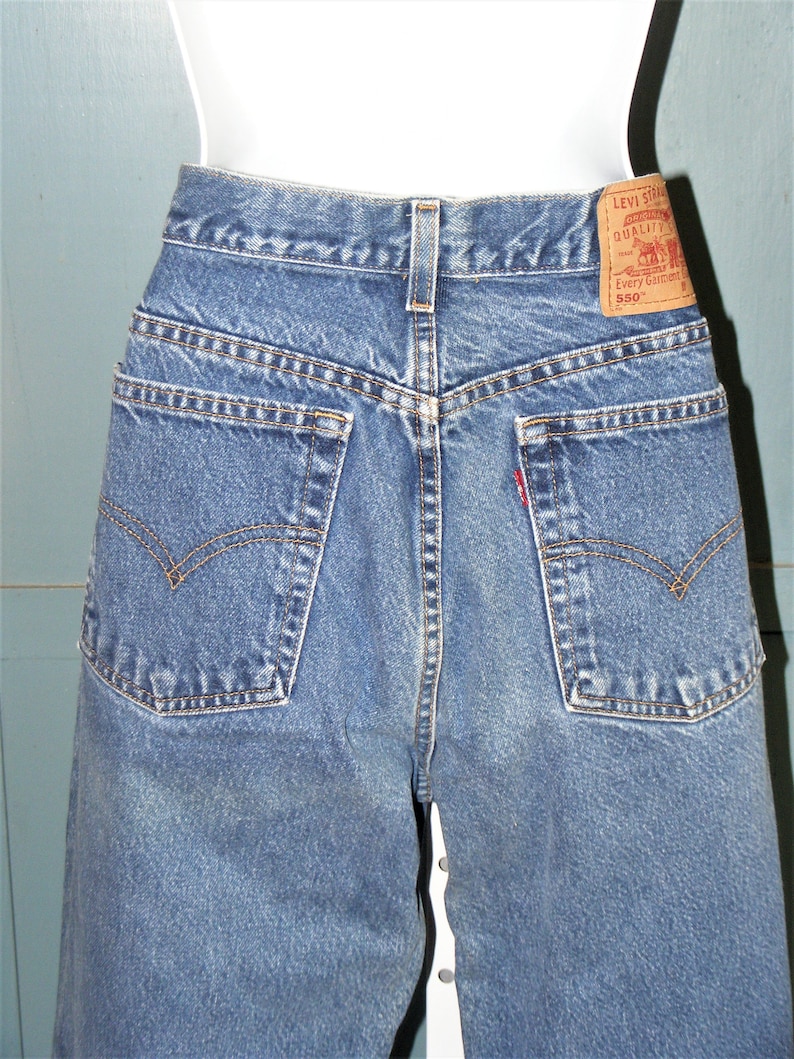 May include: Close-up of the back of blue denim jeans. The jeans have two back pockets with a classic curved design, a small red tag, and a brown leather Levi Strauss label. The jeans are displayed on a mannequin.