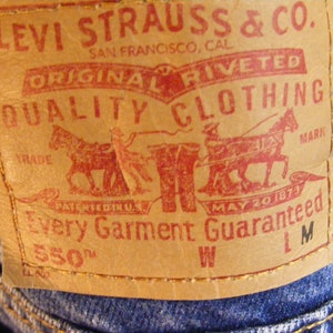 May include: Close-up of a tan leather Levi Strauss & Co. label on blue denim jeans. The red text reads "ORIGINAL RIVETED QUALITY CLOTHING" and "Every Garment Guaranteed". The label includes a graphic of two horses pulling a wagon.