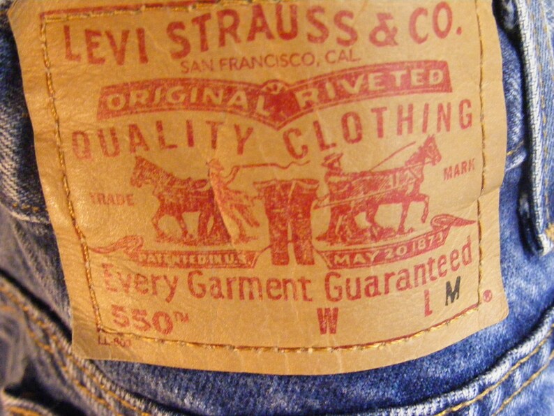 May include: Close-up of a tan leather Levi Strauss & Co. label on blue denim jeans. The red text reads "ORIGINAL RIVETED QUALITY CLOTHING" and "Every Garment Guaranteed". The label includes a graphic of two horses pulling a wagon.