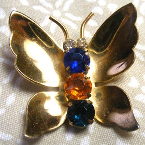 May include: A gold-toned butterfly brooch with three colorful gemstones. The brooch features a blue, orange, and teal gemstone in the center of the butterfly's body. The wings are detailed with a textured finish, and the antennae are thin and curved.