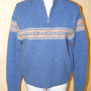 May include: A blue sweater with a half-zip collar and a patterned band across the chest and sleeves. The band features a design in cream, orange, and red. The sweater is displayed on a white mannequin.