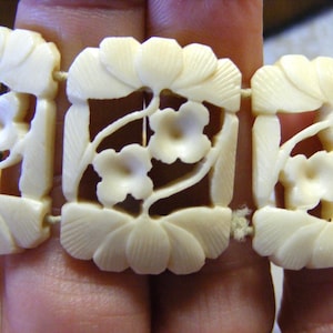Puede incluir: Pulsera de color marfil con tallados florales. La pulsera presenta eslabones rectangulares, cada uno adornado con tallados detallados de flores y hojas. Los eslabones están conectados por una cuerda, creando un diseño flexible. Primer plano.