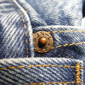 May include: Close-up of blue denim fabric with a copper-colored button. The button is embossed with the letters "LAS" and other markings. The denim has visible yellow stitching.
