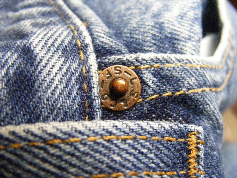 May include: Close-up of blue denim fabric with a copper-colored button. The button is embossed with the letters "LAS" and other markings. The denim has visible yellow stitching.