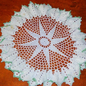 May include: A handmade, white crochet doily with a floral design. The doily features a central circular pattern surrounded by radiating petal-like shapes. The edges are trimmed with a delicate green border, adding a touch of color to the intricate lace-like design.