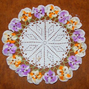 May include: A circular, handmade doily featuring a white, open-weave centre and a border of crocheted flowers in orange, purple, and white. The flowers are linked by a green crocheted vine.