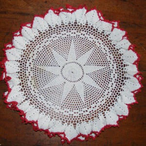 Può includere: Un centrino circolare fatto a mano, bianco con un bordo smerlato rosso. Il centrino presenta un design traforato dettagliato con un motivo a raggiera al centro. Il centrino è probabilmente realizzato in cotone o fibra simile.